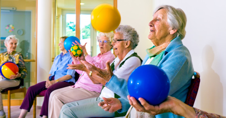 Group of happy senior ladies doing coordination exercises in a seniors gym sitting in chairs throwing and catching brightly colored balls.